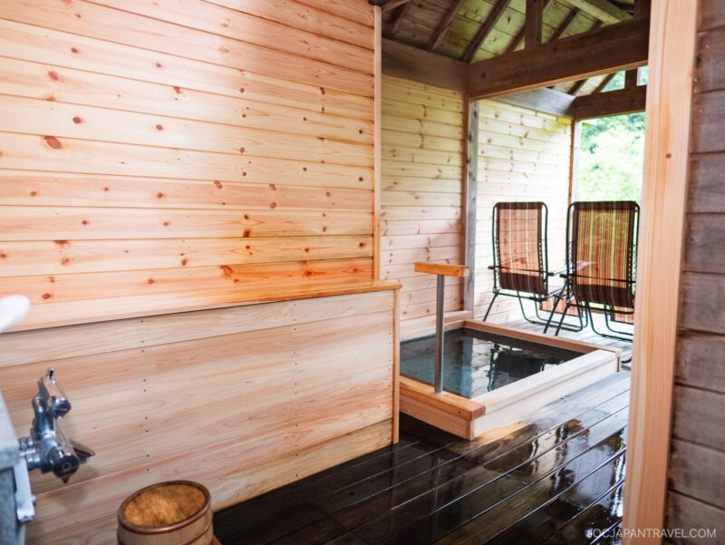 A wooden room at Nikko Okunoin Hotel Tokugawa features an indoor Japanese hot spring bath, two reclining chairs, and natural light streaming through a partially open wall, revealing lush greenery outside.