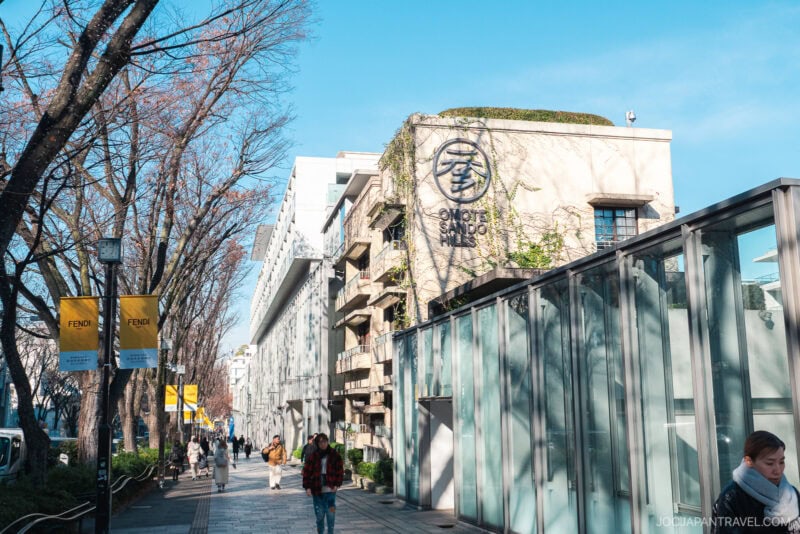 A sunny street scene in Omotesando, Tokyo, shows people strolling along a tree-lined sidewalk. Modern buildings, including one with the Omotesando Hills sign, yellow Fendi banners, and a clear blue sky complete the vibrant atmosphere.