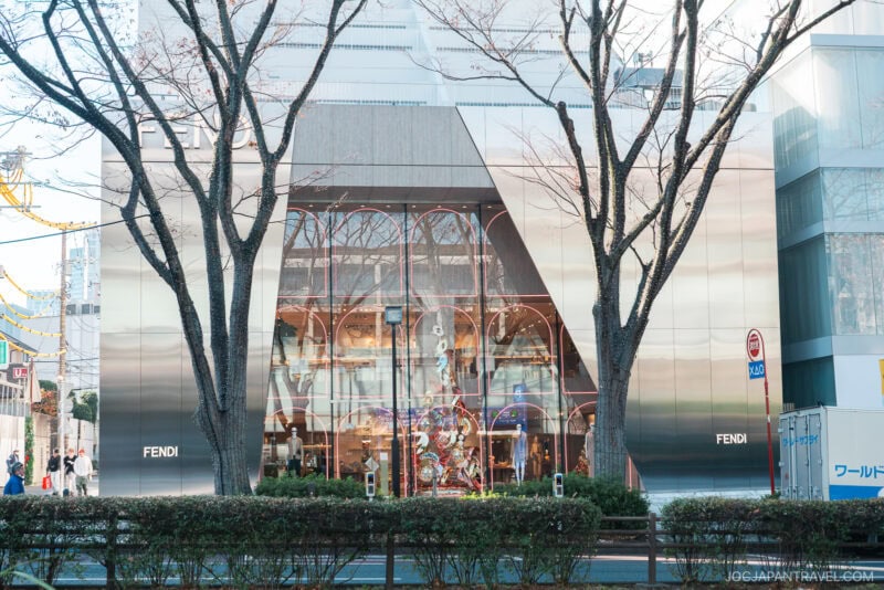 Modern Fendi store with a shiny glass and metal facade, large display windows, and bare trees in front, located on Omotesando’s city street with some greenery and construction barriers nearby.