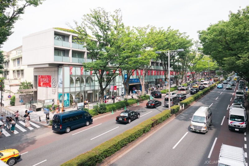 A busy Omotesando city street with cars, taxis, and vans driving past modern buildings and shops. Pedestrians stroll along the tree-lined sidewalks, and red sale banners hang from storefronts.