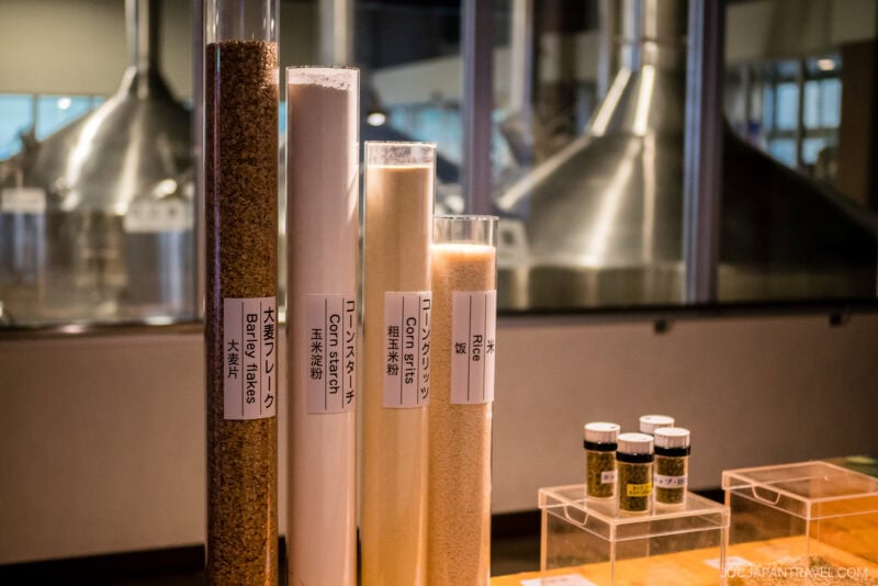 Tall cylindrical containers filled with different grains for brewing, labeled in Japanese and English, stand on a display table at Orion Happy Park. Behind them, large brewing equipment is visible through glass windows.
