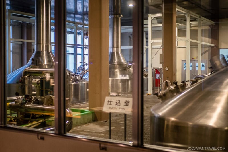View through glass of Orion Happy Park’s modern brewery with large stainless steel tanks and pipes, a sign labeled Mash Pan in English and Japanese, and clean, industrial surroundings.