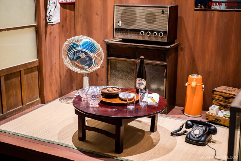 A retro Japanese room with a round table set with glasses, snacks, and a beer bottle—perhaps from Orion Happy Park—an old radio, vintage fan, orange thermos, rotary phone, and tatami mats on the floor.