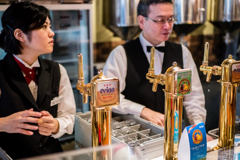 Two bartenders in uniforms stand behind a bar with gold Orion beer taps at Orion Happy Park. Glassware and drink menus are on the counter, set against a sleek, modern bar backdrop.