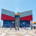 Osaka Aquarium Kaiyukan (海遊館) – Whale Sharks and Pacific Wonders Visitors stroll near the vibrant, modern facade of Osaka Aquarium Kaiyukan in Japan, with its large glass panels, sea creature mural, and striking central structure glowing under a clear blue sky.