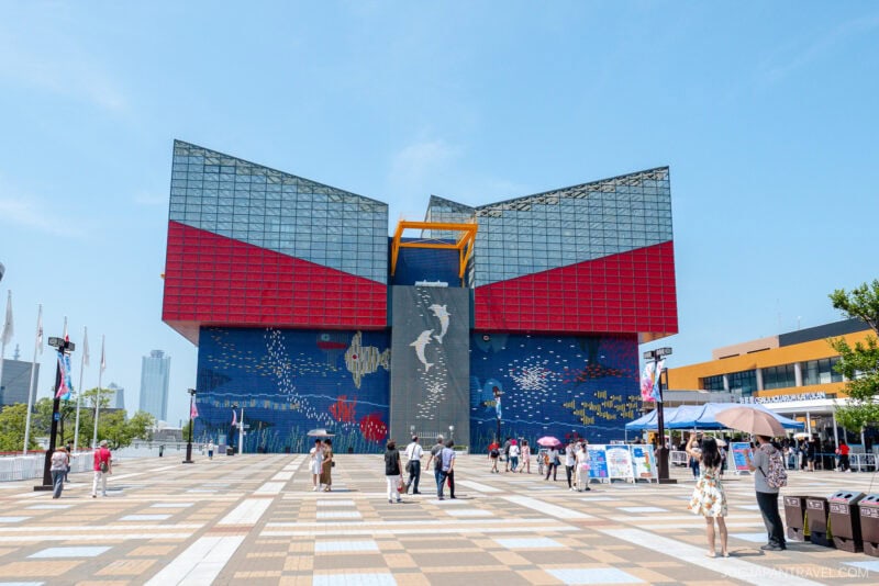 Visitors stroll near the vibrant, modern facade of Osaka Aquarium Kaiyukan in Japan, with its large glass panels, sea creature mural, and striking central structure glowing under a clear blue sky.
