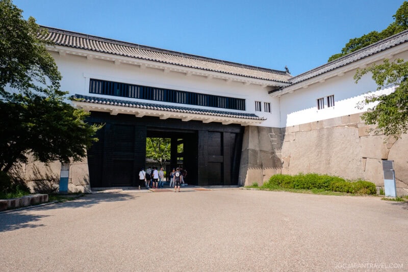 A group of people walk toward the large gate of Osaka Castle, featuring white walls, black wooden doors, and a sturdy stone foundation, surrounded by trees on a sunny day.
