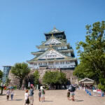 Exploring Osaka Castle (大阪城) Osaka Castle shines on a sunny day, surrounded by lush trees and visitors enjoying the spacious plaza in front of the iconic white and green Osaka Castle.
