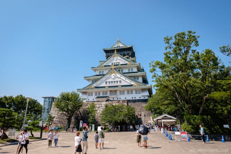 Osaka Castle shines on a sunny day, surrounded by lush trees and visitors enjoying the spacious plaza in front of the iconic white and green Osaka Castle.