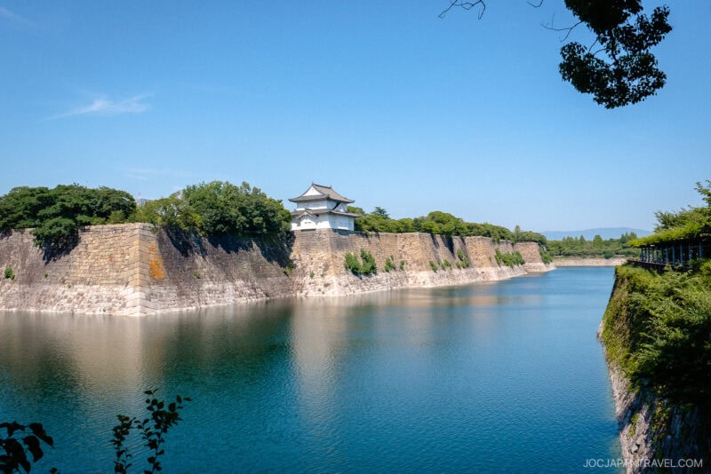A white traditional Japanese castle turret, part of Osaka Castle, sits atop tall stone walls, surrounded by a calm blue moat and lush green trees under a clear blue sky.