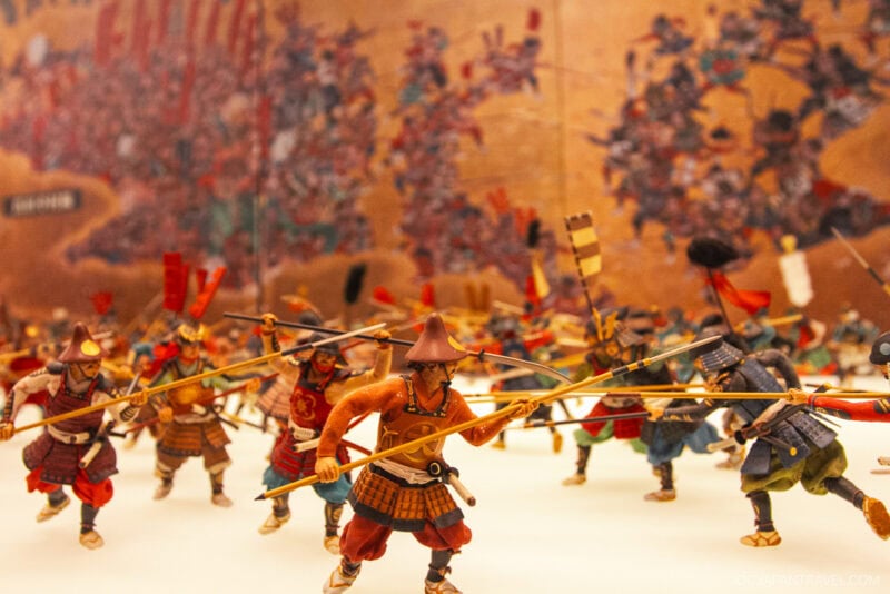 Miniature samurai warriors in traditional armor, holding spears and banners, are arranged in dynamic battle poses before a detailed painted backdrop depicting a historic battle scene near Osaka Castle.