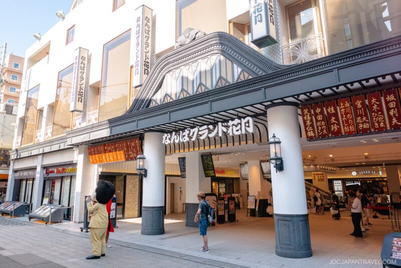 The entrance to Namba Grand Kagetsu theater in Namba Osaka features white columns, Japanese signage, and people walking by—including someone in a bear costume near the entrance.