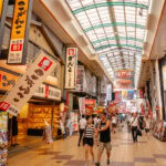 A busy indoor shopping street in Namba, Osaka, with colorful signs in Japanese, shops on both sides, and people walking along the tiled walkway under a glass roof.