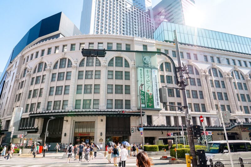 A busy Osaka Namba city street with pedestrians crossing in front of a large, white, multi-story building with arched windows; modern skyscrapers rise in the background under a sunny sky.