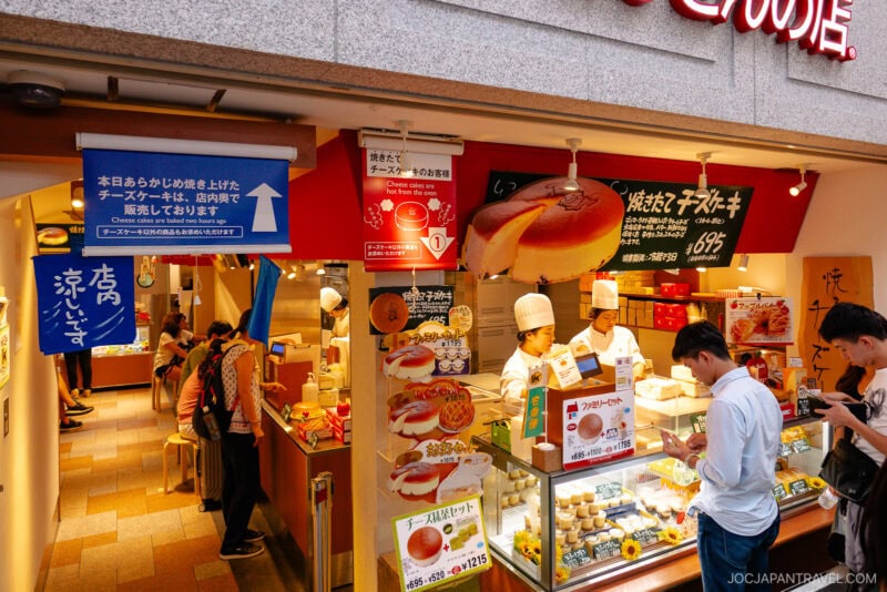A bakery shop in Osaka Namba, Japan, with chefs preparing and selling cheesecakes behind a counter, while customers wait and browse. The colorful storefront features signs and menus displayed in Japanese.