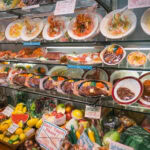 Display case filled with realistic Plastic Food Sampuru models—pasta, steaks, desserts, and fruits—arranged on plates and trays with signs reading “Don’t Touch Please” in a restaurant or shop setting.