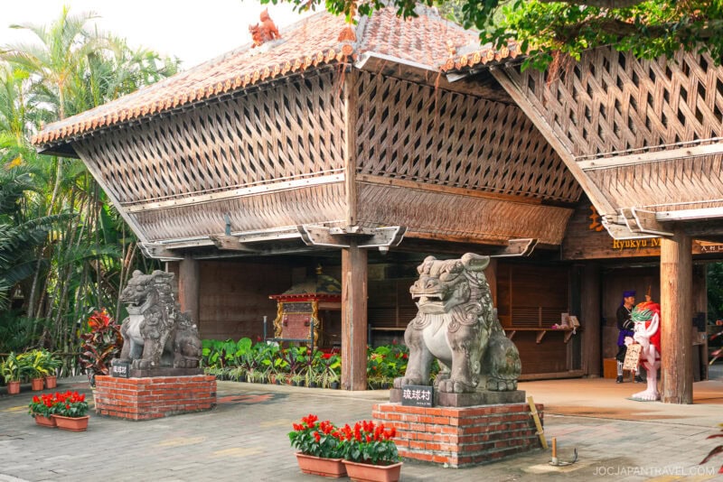 Traditional Okinawan building with a tiled roof, wooden walls, and two large stone Shisa lion-dog statues at the entrance, surrounded by plants and flowers in the heart of Ryuku Mura.