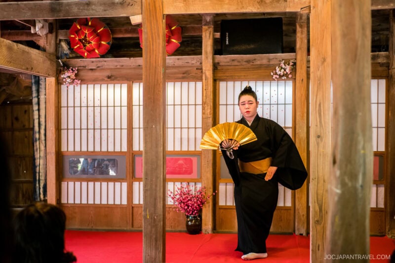 A performer in a black kimono holds an open gold fan while posing on a red-carpeted stage at Ryuku Mura, a traditional Japanese wooden room adorned with red lanterns and flowers.