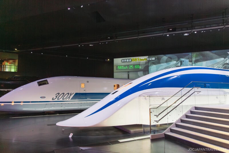 Two futuristic high-speed trains, including the white and blue Maglev and the 300X Shinkansen, are displayed indoors at SCMaglev and Railway Park under spotlights, with a staircase in the foreground and a digital sign in the background.