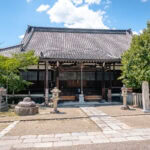 Traditional Nara Saidai-ji wooden temple with a tiled roof, surrounded by greenery and stone lanterns, under a bright blue sky with scattered clouds. A stone path leads to the entrance.