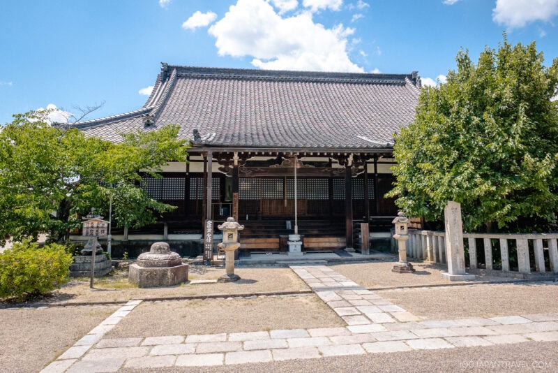 Traditional Nara Saidai-ji wooden temple with a tiled roof, surrounded by greenery and stone lanterns, under a bright blue sky with scattered clouds. A stone path leads to the entrance.