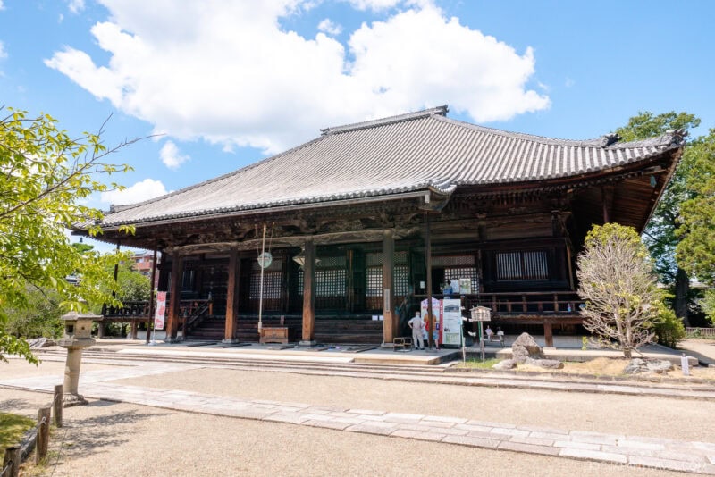 A traditional Japanese wooden temple, Nara Saidai-ji, with a tiled roof and open veranda stands under a blue sky with scattered clouds, surrounded by trees and a stone lantern in the foreground.
