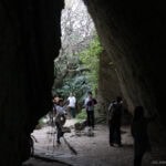 A group of people explore a large, rocky cave at Seifa-Utaki, with light streaming in from outside and greenery framing the entrance. Some are taking photos while others admire the sacred site.