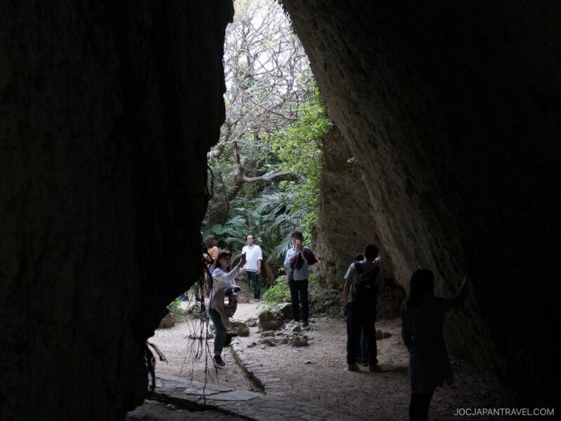 A group of people explore a large, rocky cave at Seifa-Utaki, with light streaming in from outside and greenery framing the entrance. Some are taking photos while others admire the sacred site.