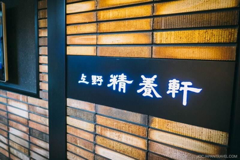 A close-up of a brick wall with illuminated Japanese characters displayed on a black panel. The Seiyoken Ueno sign glows brightly in white and blue, contrasting with the warm tones of the brick background.