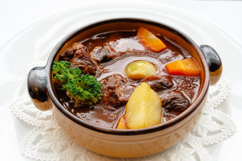 A brown ceramic bowl filled with Seiyoken beef stew, featuring chunks of meat, potatoes, carrots, broccoli, and a pearl onion in a rich, dark broth, placed on a white lace doily.