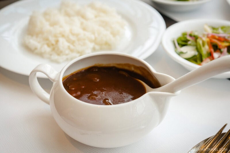 A white gravy boat filled with rich brown stew from Seiyoken Ueno sits next to a plate of plain white rice and a bowl of mixed salad on a crisp white tablecloth.
