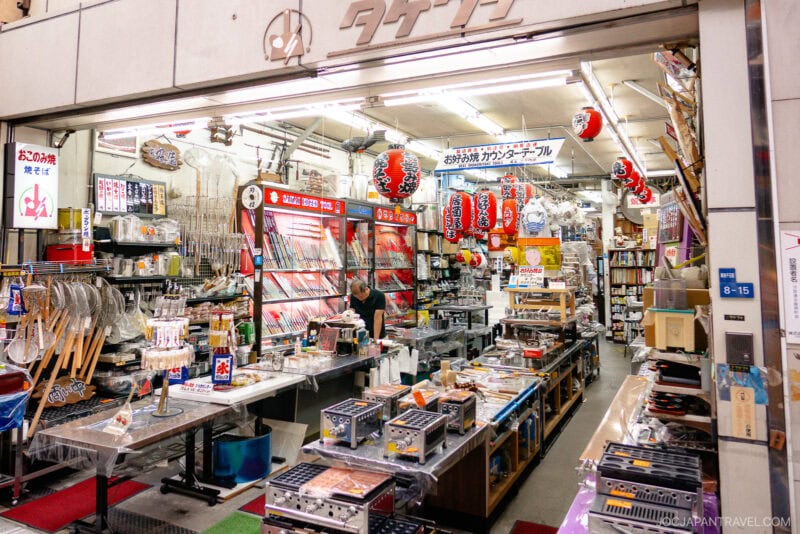 A brightly lit Japanese kitchenware shop on Sennichimae Kitchenware Street, filled with utensils, knives, grills, and red lanterns, with a person arranging items behind the counter.