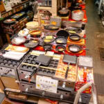 Sennichimae Kitchenware Street (千日前道具屋筋) A shop display table on Sennichimae Kitchenware Street filled with various Japanese cooking pans, molds, and utensils—including takoyaki pans and grills—with price tags and packaging visible in a busy store setting.