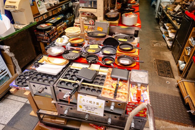 A shop display table on Sennichimae Kitchenware Street filled with various Japanese cooking pans, molds, and utensils&mdash;including takoyaki pans and grills&mdash;with price tags and packaging visible in a busy store setting.