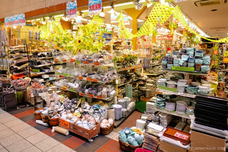 A colorful Japanese store on Sennichimae Kitchenware Street, filled with neatly arranged plates, bowls, and utensils, features bright decorations hanging from the ceiling and price tags on many vibrant kitchen items.