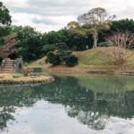 Shikinaen (識名園) – The Royal Garden of the Ryukyu Kings A tranquil Shikinaen park scene with a stone bridge over a reflective pond, grassy mounds, trees with sparse leaves, and a cloudy sky in the background.