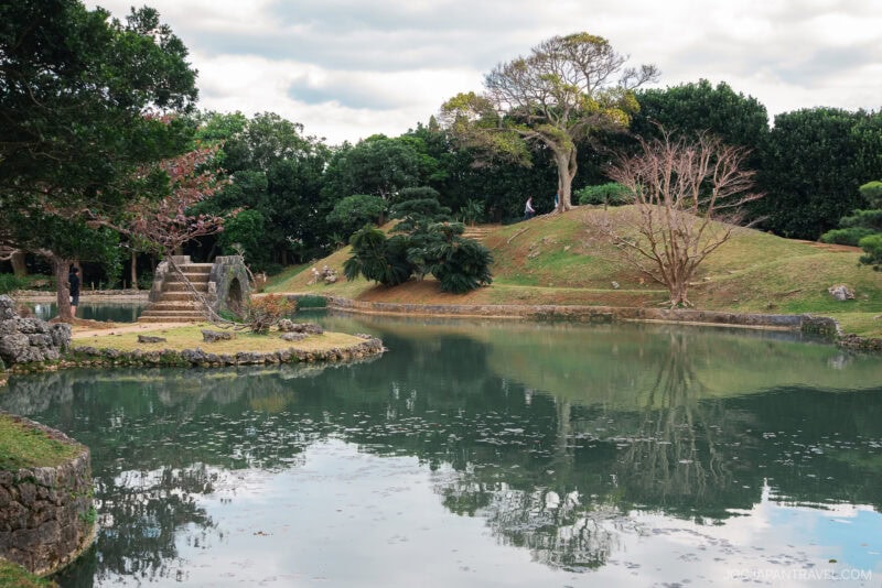 A tranquil Shikinaen park scene with a stone bridge over a reflective pond, grassy mounds, trees with sparse leaves, and a cloudy sky in the background.