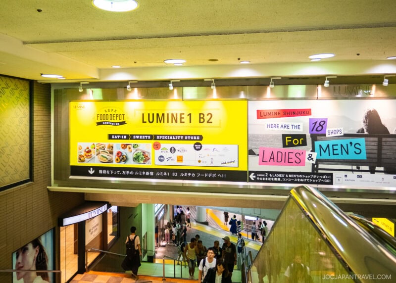 An escalator in Shinjuku descends into an underground shopping area, where large yellow signs advertise LUMINE 1 B2 and 18 shops for ladies & men’s, featuring food depictions and directional arrows.