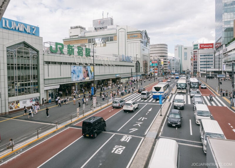 A busy Shinjuku street scene outside Shinjuku Station in Tokyo, Japan, with cars, buses, and pedestrians. Buildings with signs like Lumine 2 and JR line the street under a partly cloudy sky.