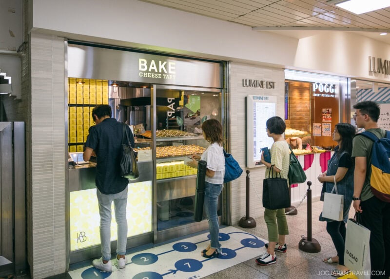 Customers stand in line at Bake Cheese Tart, a small shop in Shinjukus brightly lit indoor space, waiting to purchase baked goods. Boxes of products are stacked behind the counter.