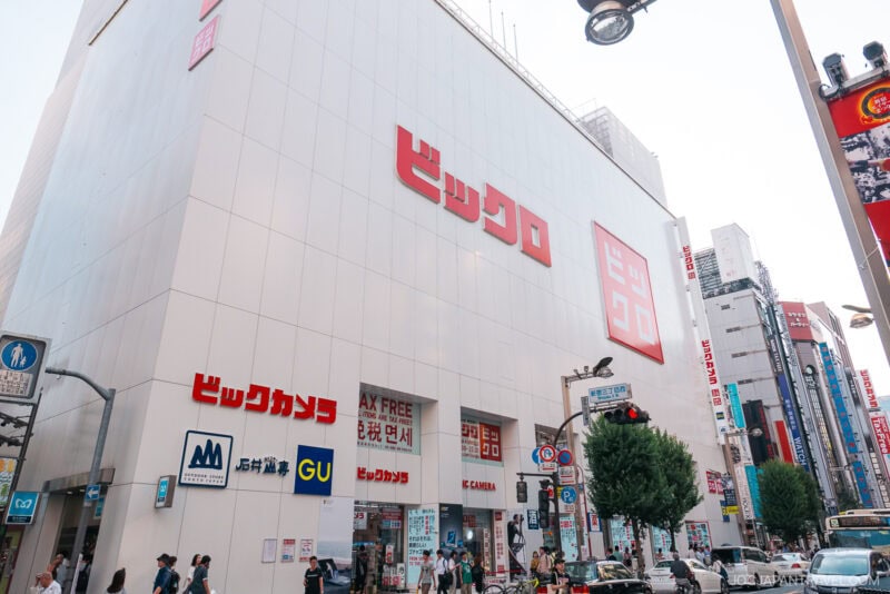 A large white building in Shinjuku with bold red Japanese signage and several shop logos at street level. People walk on the sidewalk and cars drive by under a clear sky.