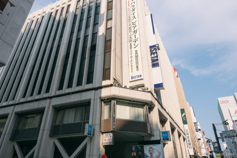 A tall, modern white building with ISETAN signage rises in Shinjuku, featuring large vertical banners with Japanese text, glass windows, and blue sky above. Pedestrians and nearby signs are visible at street level.