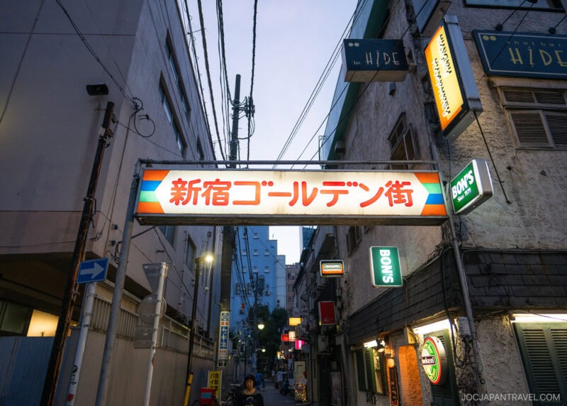 A street entrance in Shinjuku Golden Gai, marked by a colorful sign with Japanese characters, is surrounded by narrow buildings, neon lights, and overhead wires glowing at dusk.
