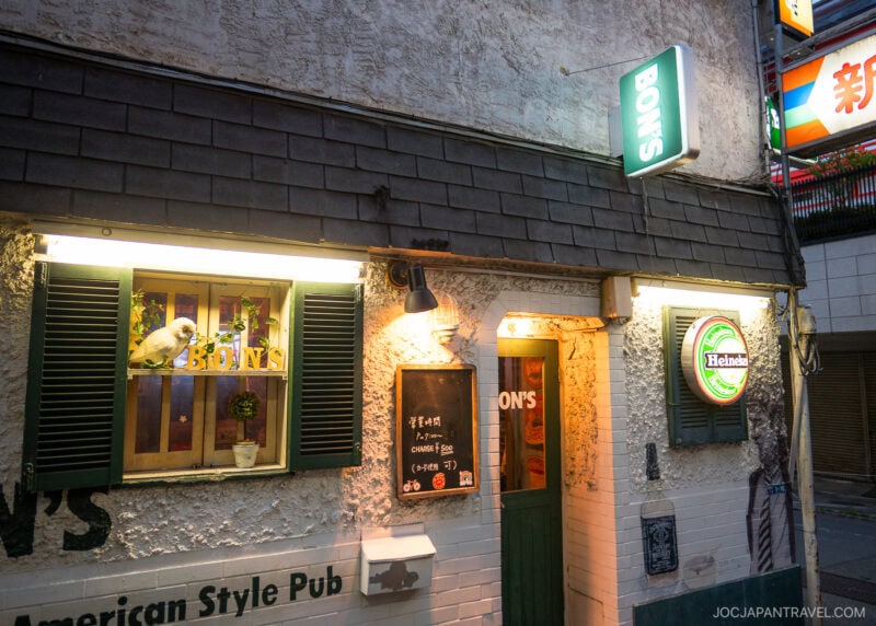 A white parrot sits in a window of BONS, an American-style pub with a green door, chalkboard menu, and Heineken sign, illuminated by warm exterior lights in Shinjuku Golden Gais urban Japan setting.