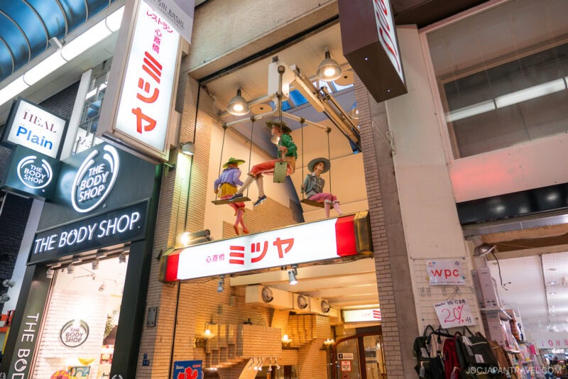 A storefront in Osaka Shinsaibashi with a sign reading ミツヤ (Mitsuya) features three mannequins dressed as musicians playing instruments on a swing above the entrance, next to a Body Shop store.