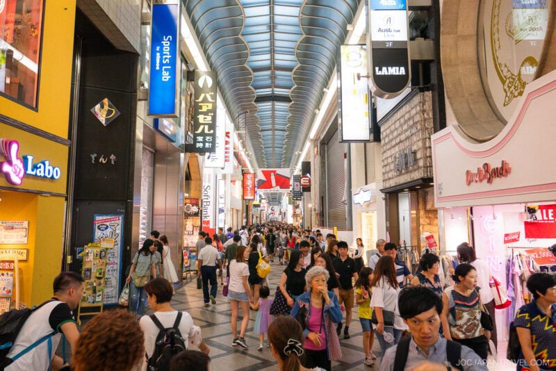 A busy indoor shopping street in Osaka Shinsaibashi, filled with people walking and browsing stores; colorful shop signs and advertisements line both sides under a curved glass roof.