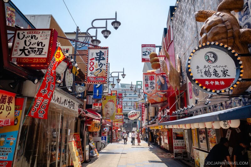 A vibrant Japanese street in Shinsekai Osaka, lined with colorful signs, lanterns, and restaurants—some boasting large food displays and playful mascots. A few people stroll the lively pathway beneath a bright blue sky.