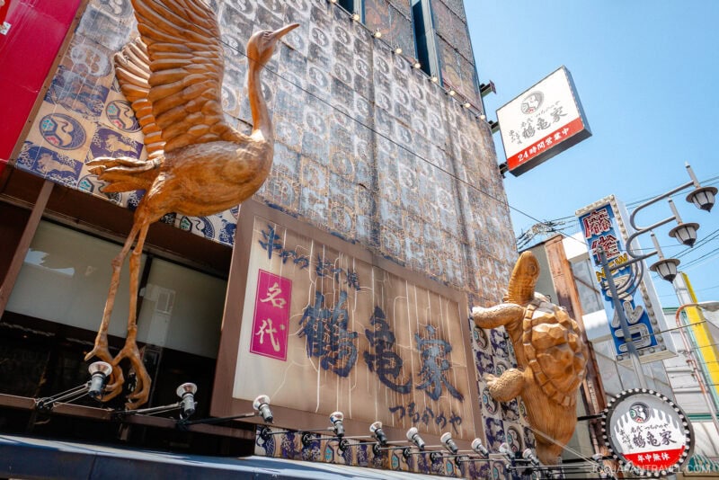 Large golden crane and turtle statues adorn the exterior of a Japanese building in Shinsekai Osaka, surrounded by vibrant signs with Japanese characters under a bright blue sky.