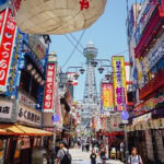 Shinsekai (新世界) – Osaka’s Retro New World A bustling street in Shinsekai Osaka, lined with colorful storefronts and bright signs in Japanese, with Tsutenkaku Tower rising in the background and many people walking below.