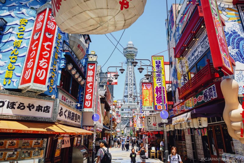 A bustling street in Shinsekai Osaka, lined with colorful storefronts and bright signs in Japanese, with Tsutenkaku Tower rising in the background and many people walking below.